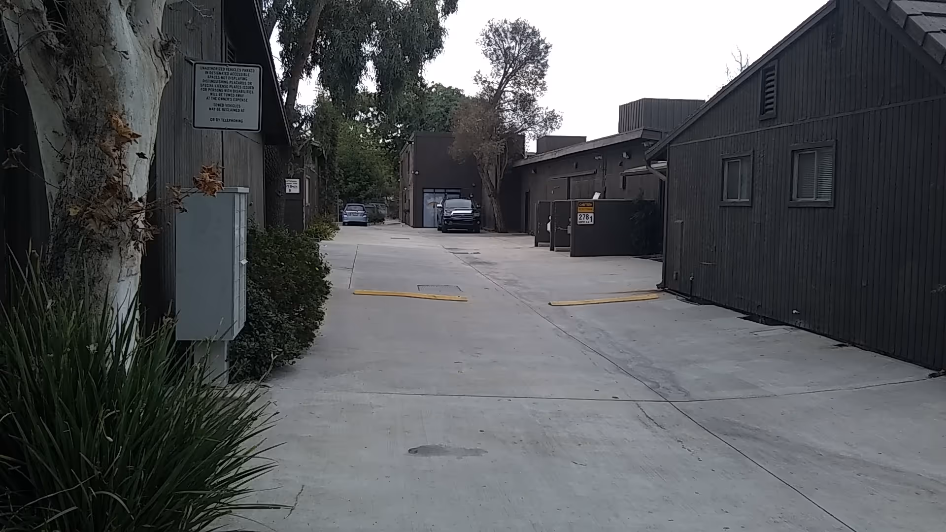 A concrete alley between dark wooden buildings with dumpsters and parked cars.