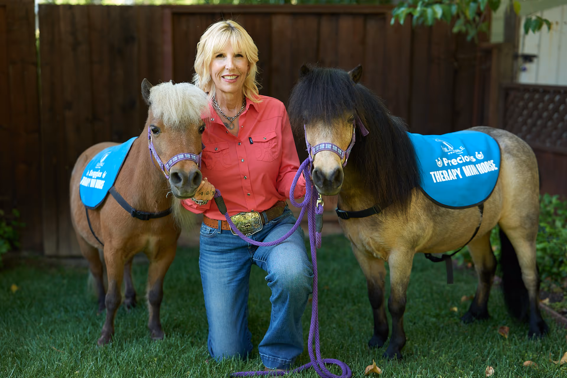 Smiling woman kneels with two ponies on a grassy outdoor yard.