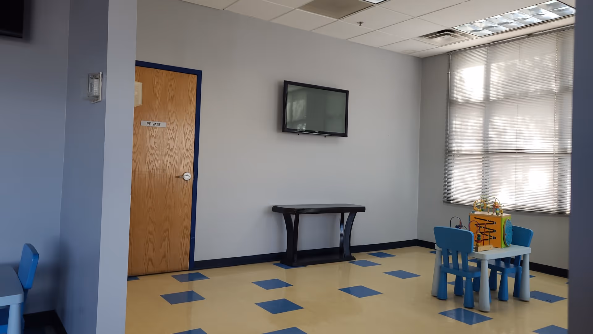An indoor common area with a wall-mounted TV, a wooden door, and blue children's play table.