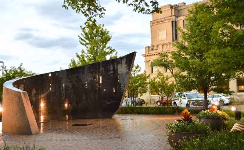 Outdoor courtyard with a curved black stone fountain, trees, planters, and a building in background