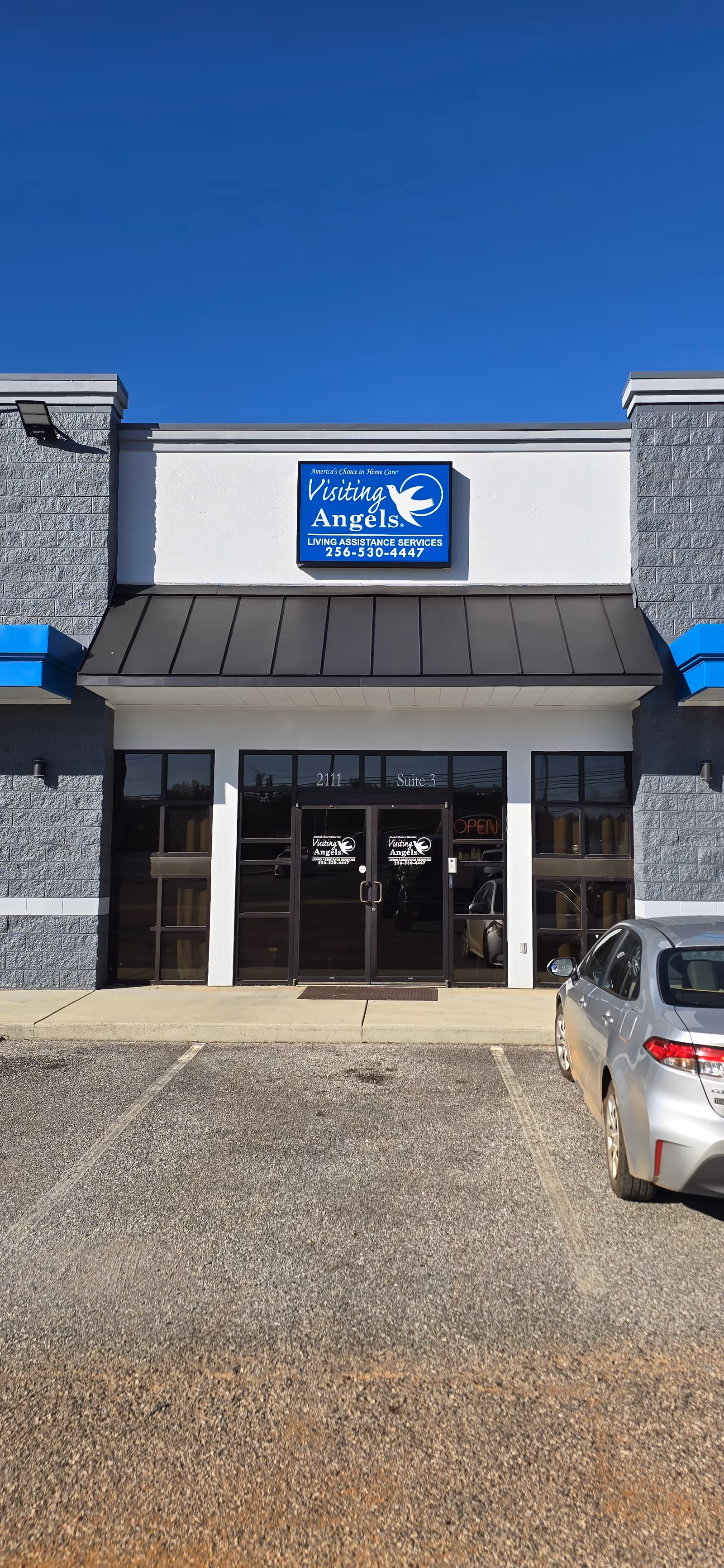 Front view of Visiting Angels storefront with blue sign, glass doors, and a parked car.