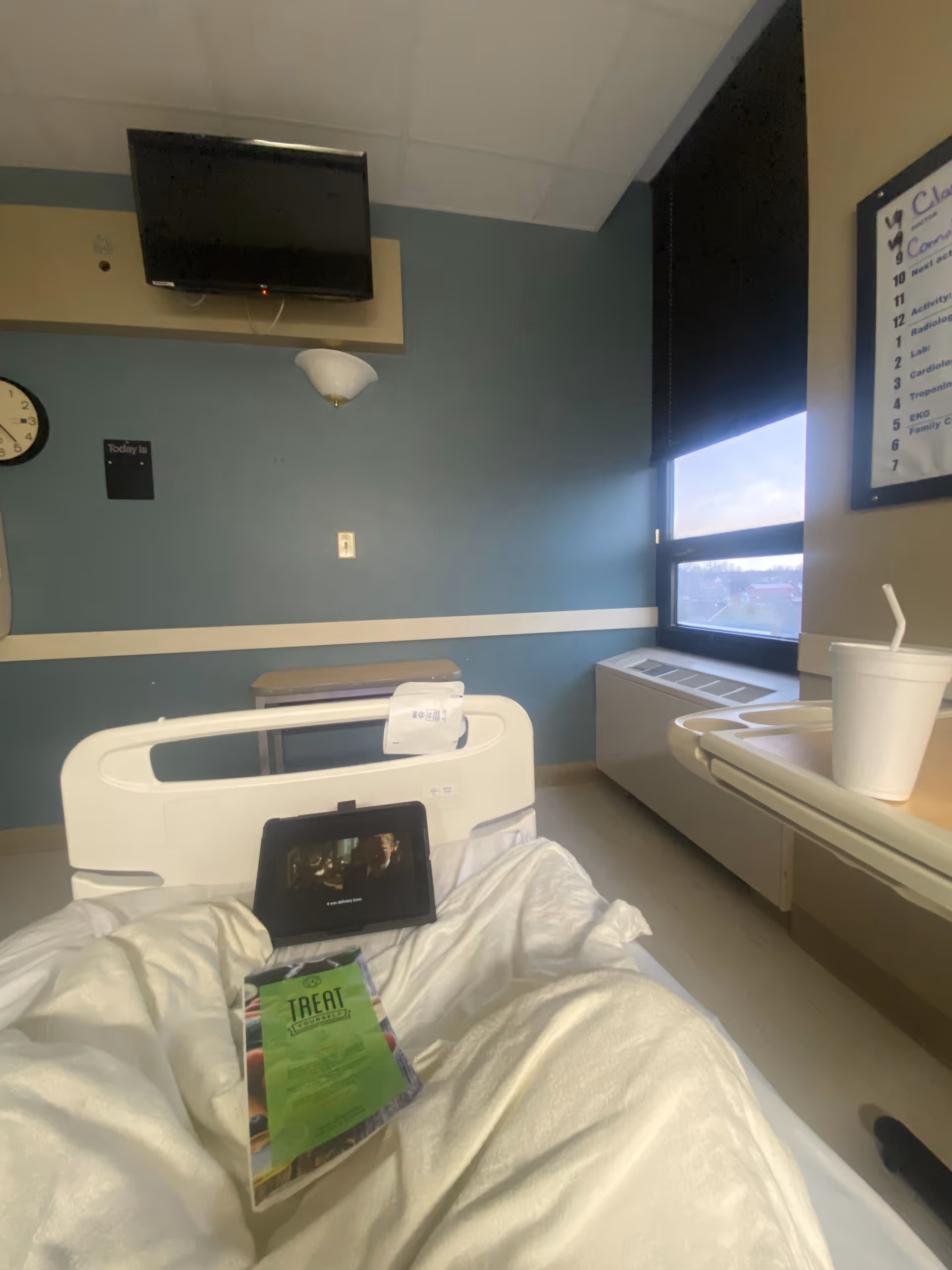 Hospital room with a bed, wall-mounted TV, window, and a tablet on the bed.