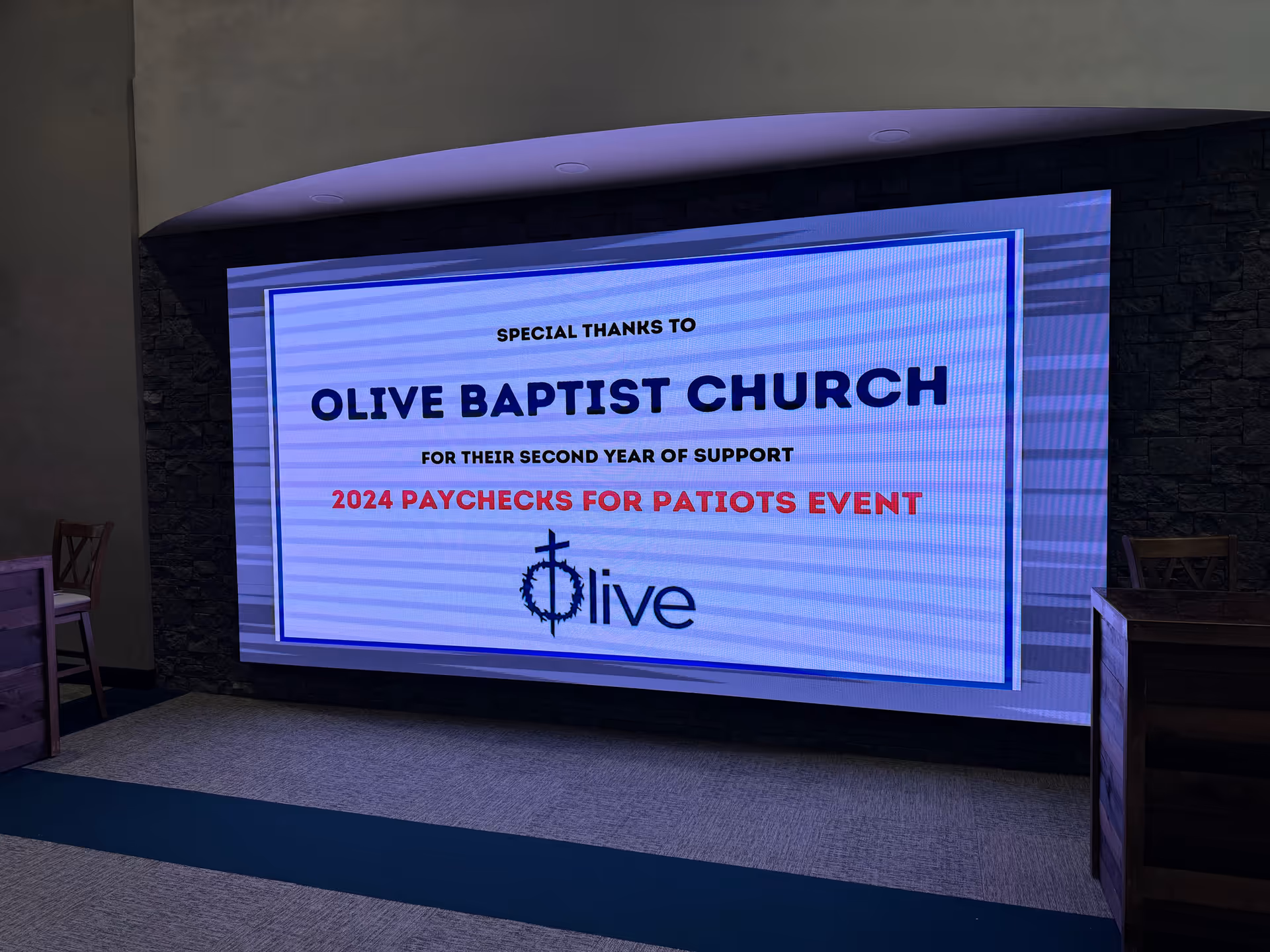 Large digital screen in a dim interior shows a thank-you message to Olive Baptist Church