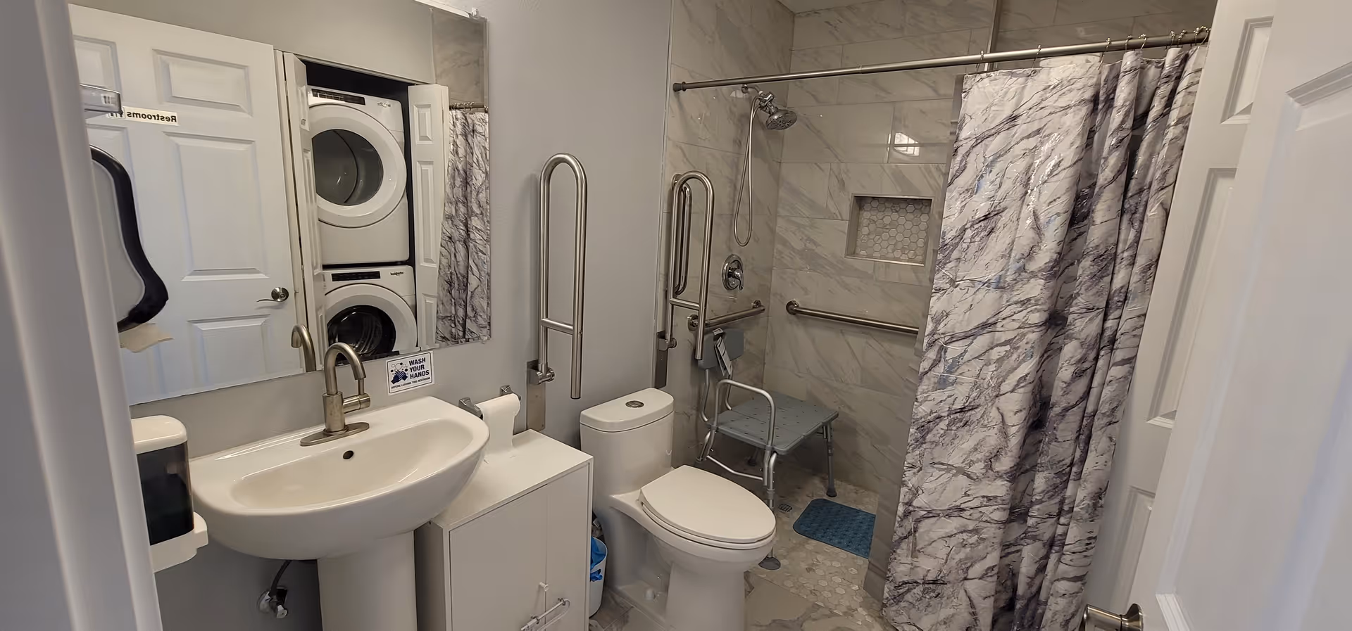 Accessible bathroom with sink, toilet, shower with grab bars, a shower chair, and stacked laundry appliances.