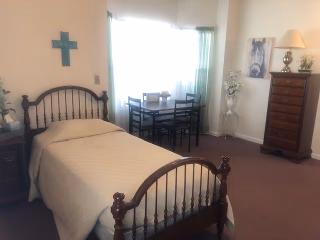 A bedroom with a wooden bed, dresser, and dining table by a bright window.