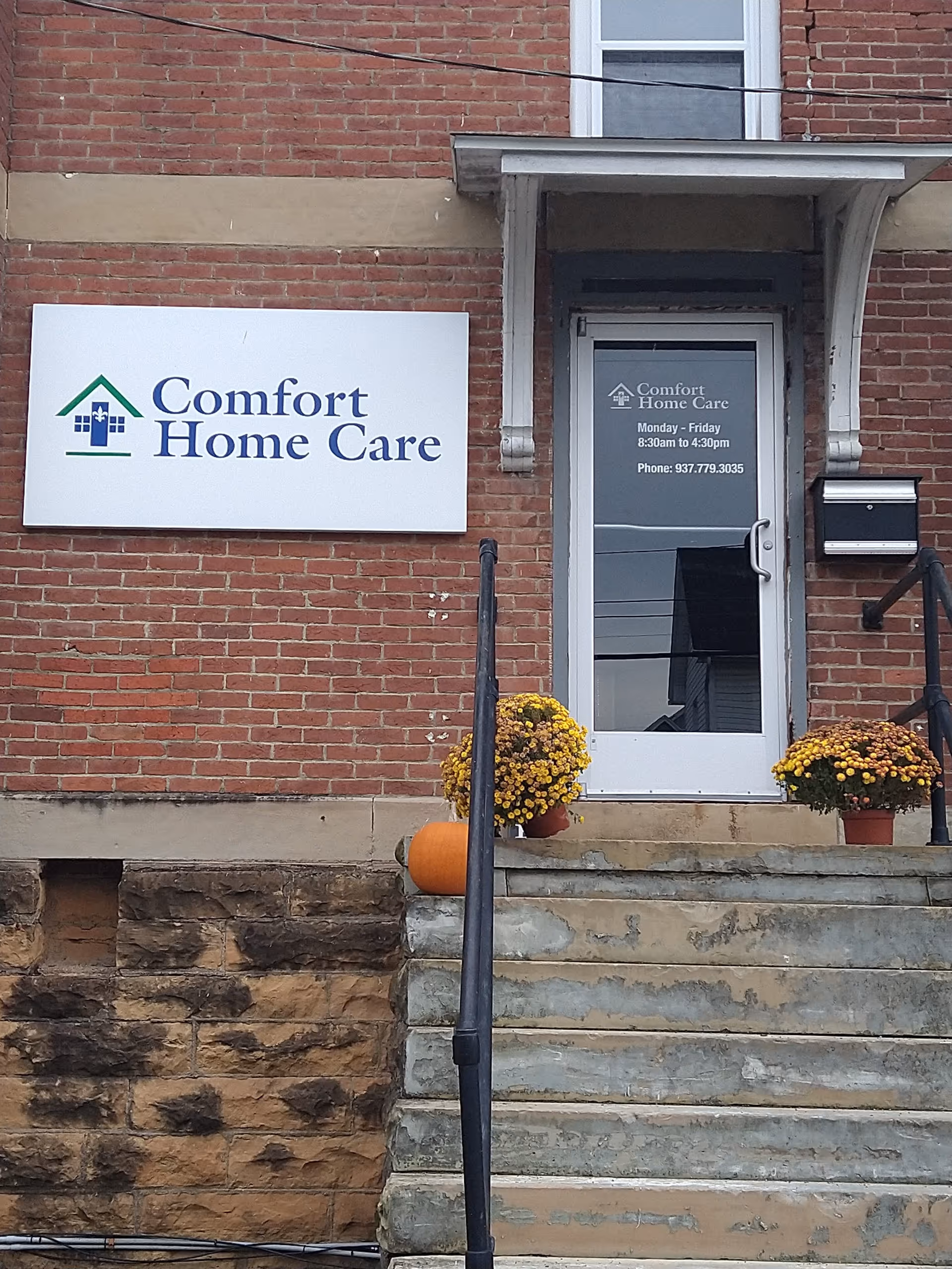 Brick building entrance for Comfort Home Care with sign, stairs, railing, and pumpkins.