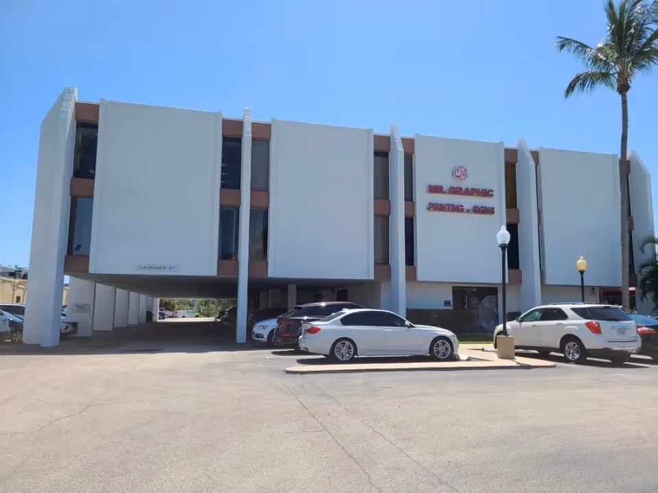 Exterior view of a white multi-story building with parked cars and palm trees.