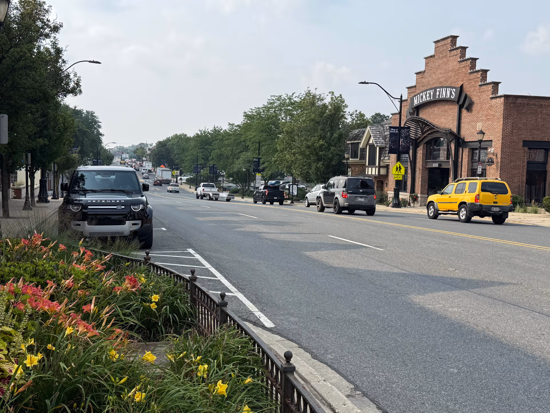 Street view with parked cars, blooming flowers, and a brick building labeled Mickey Finn's.