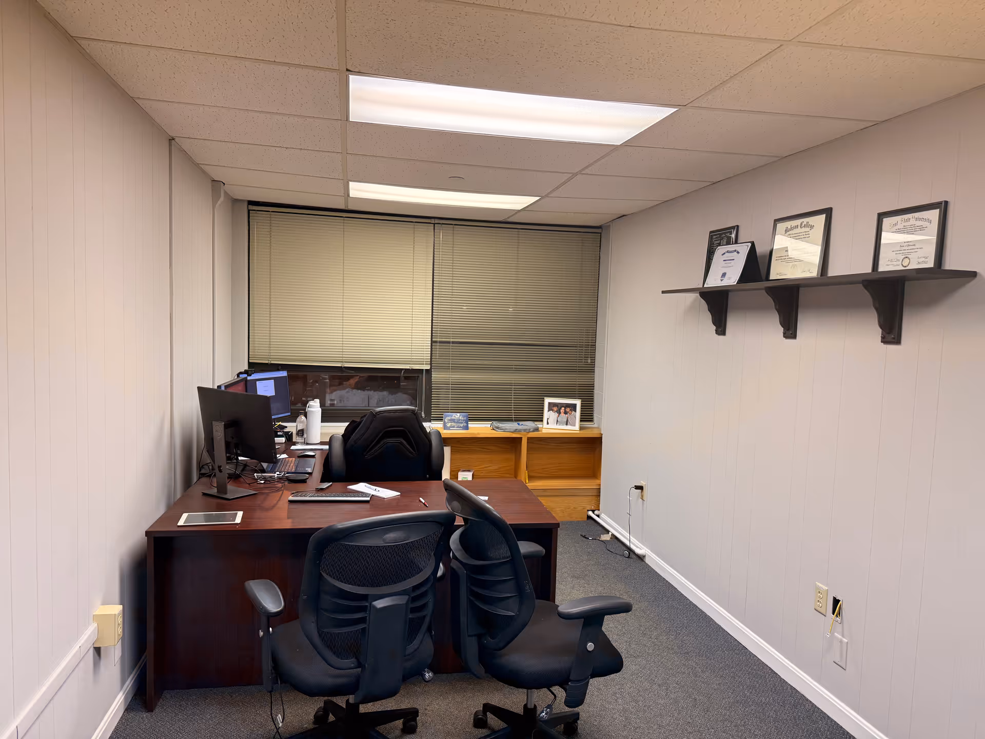 Small office with desk, computer, two chairs, blinds, and framed certificates on wall.