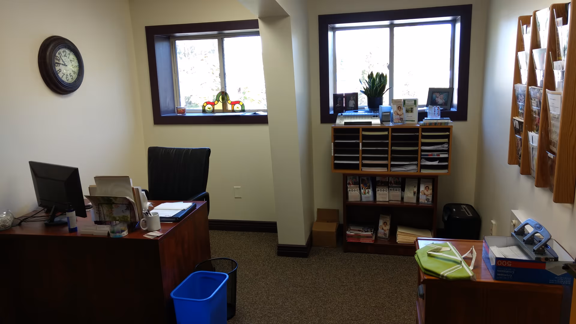 Office reception area with desk, computer, filing shelves, window light, and magazine rack.