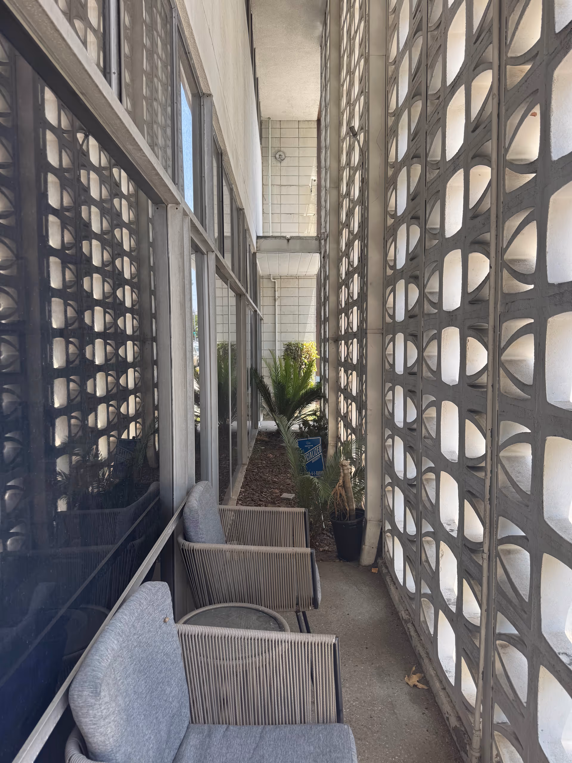Narrow outdoor corridor with lattice walls, chairs, and potted plants along the building.