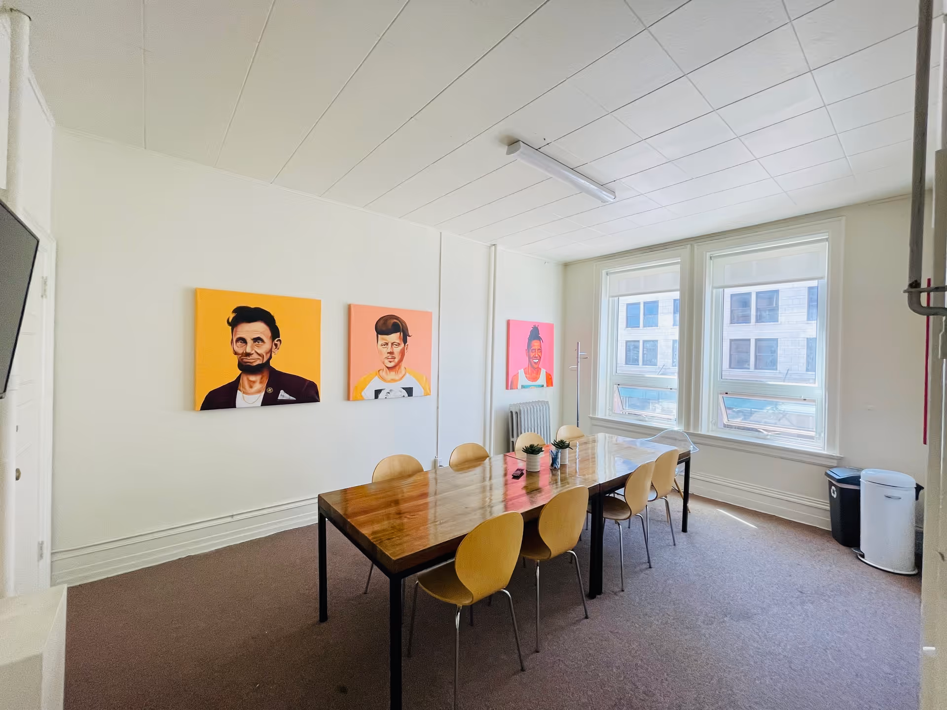 Bright dining room with long wooden table, yellow chairs, and colorful portraits on the wall.