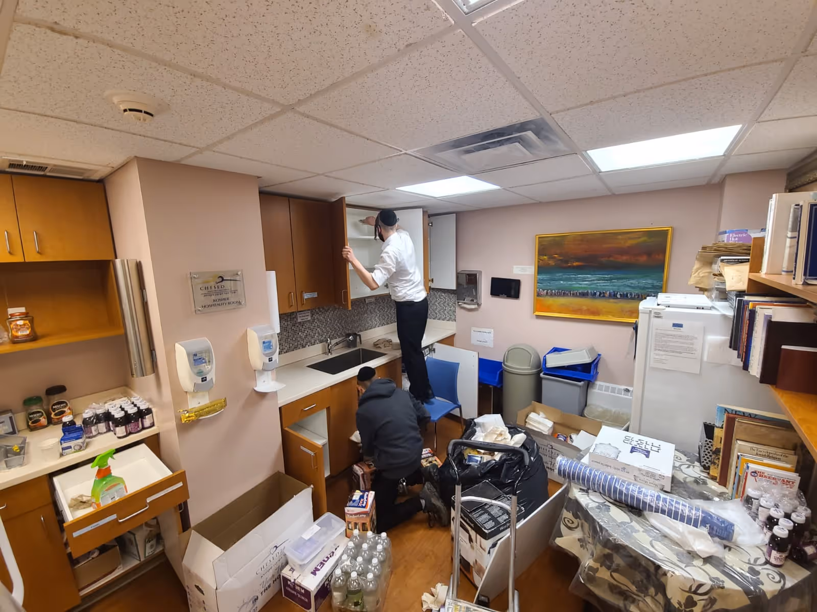 Two workers in a cluttered staff kitchen area sorting supplies.