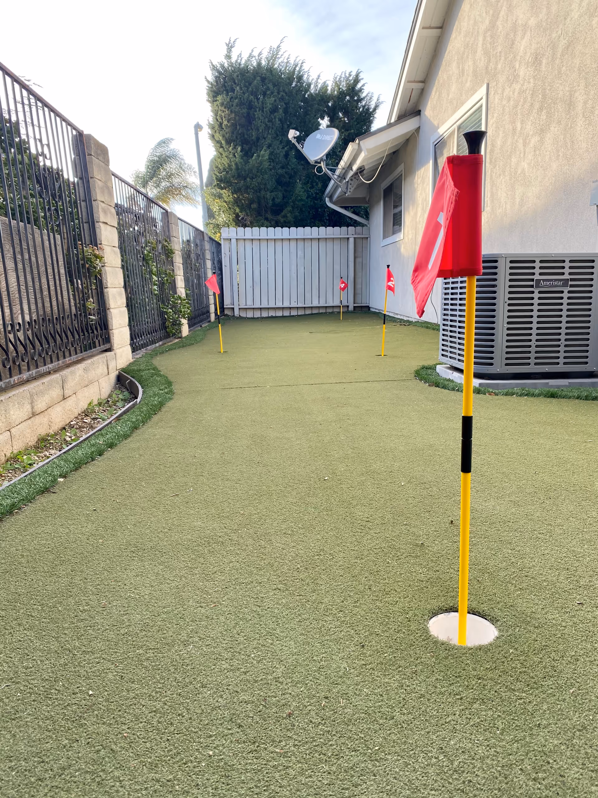 Outdoor courtyard with artificial turf, flag poles, fence, satellite dish, and building air conditioner.