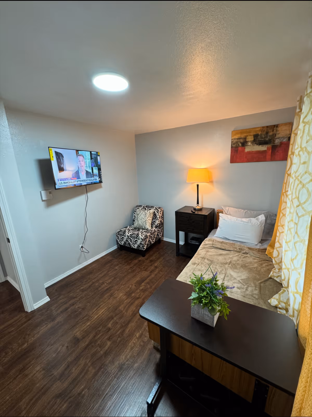 Cozy furnished bedroom with bed, nightstand, chair, wall TV, and a small plant.