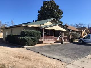 Single-story building with a covered porch, green roof, shrubs, and a parked vehicle.