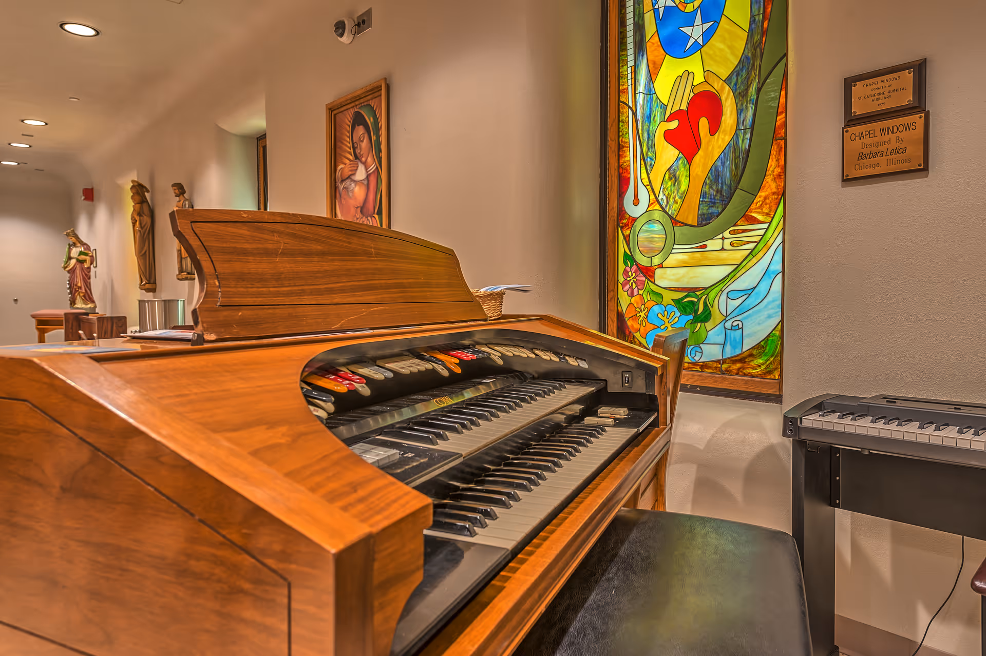 Wooden piano/organ in a hallway with religious statues and a stained-glass panel.