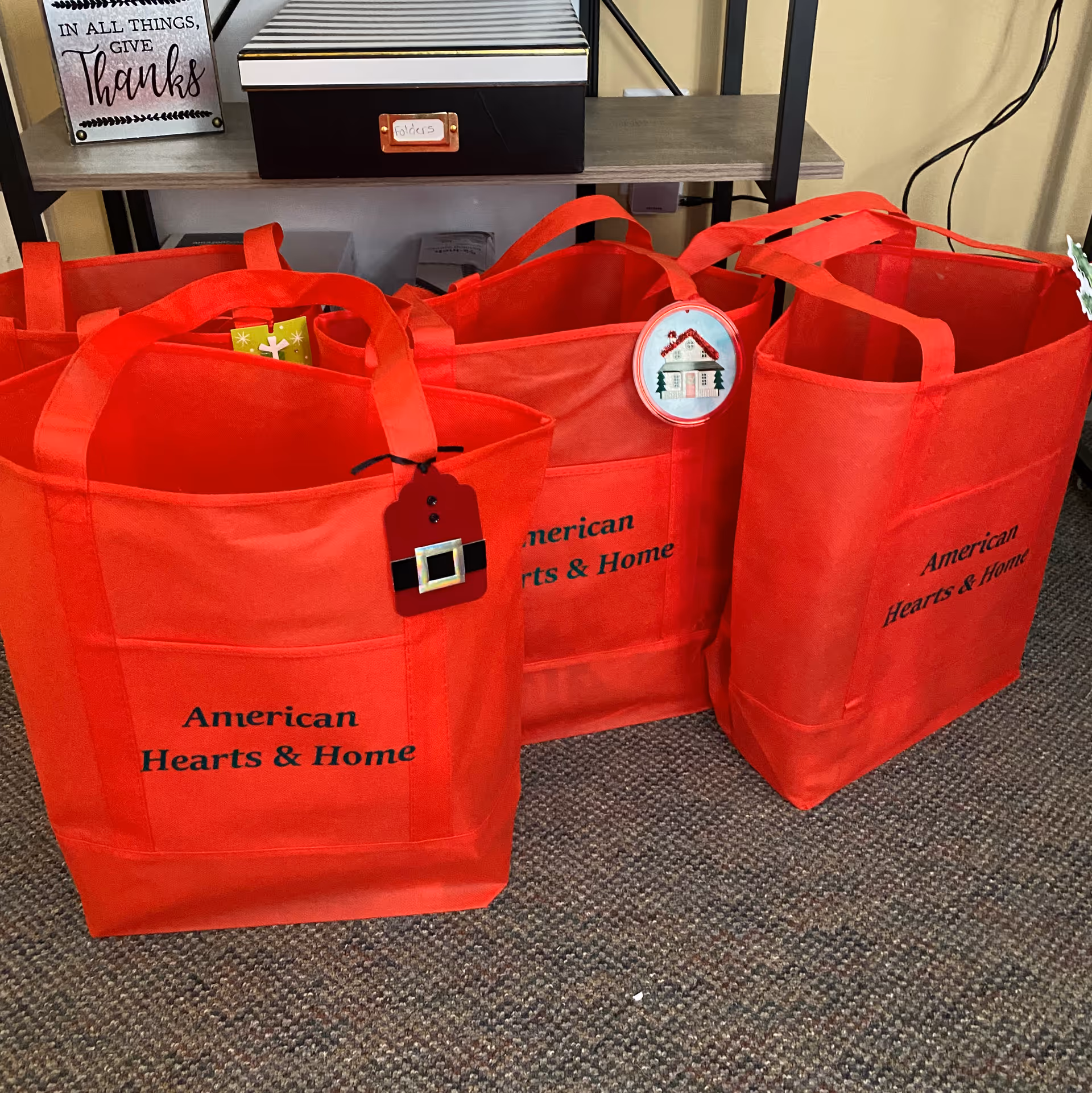 Red tote bags labeled American Hearts & Home sit on carpet beside shelves indoors.