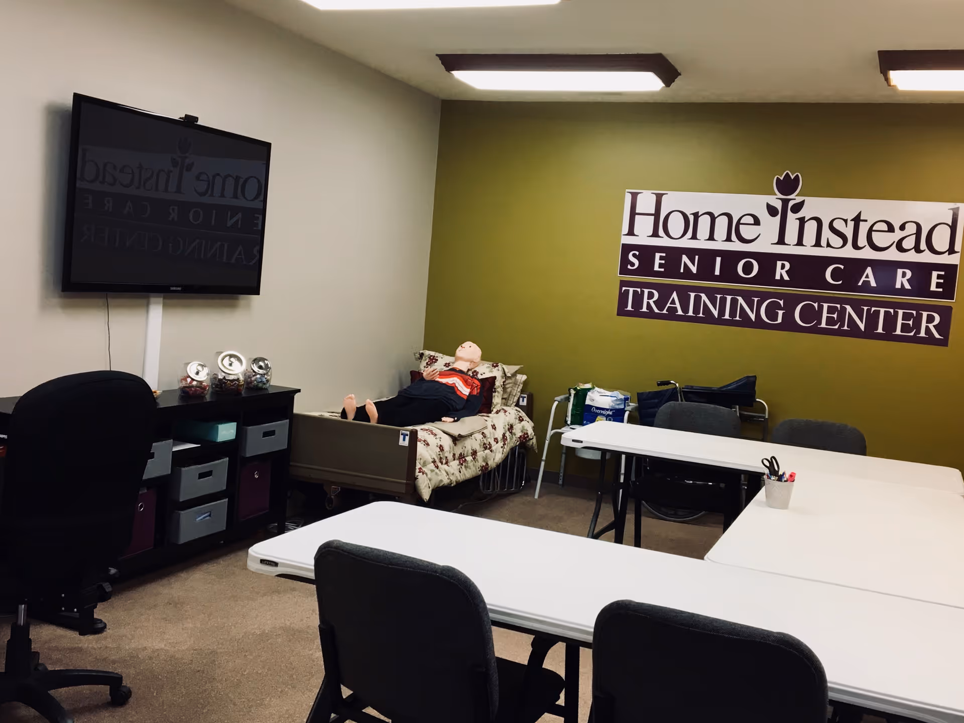 A training room inside Home Instead Senior Care with tables, chairs, and a mannequin on bed.