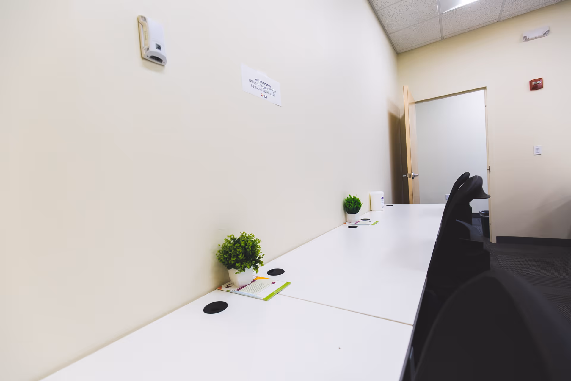 Long white counter with small green plants and black chairs along a bright interior wall.