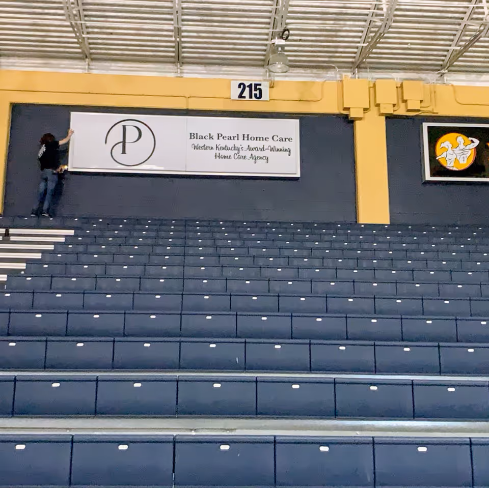 Two people stand on blue stadium bleachers beside a wall sign reading Black Pearl Home Care