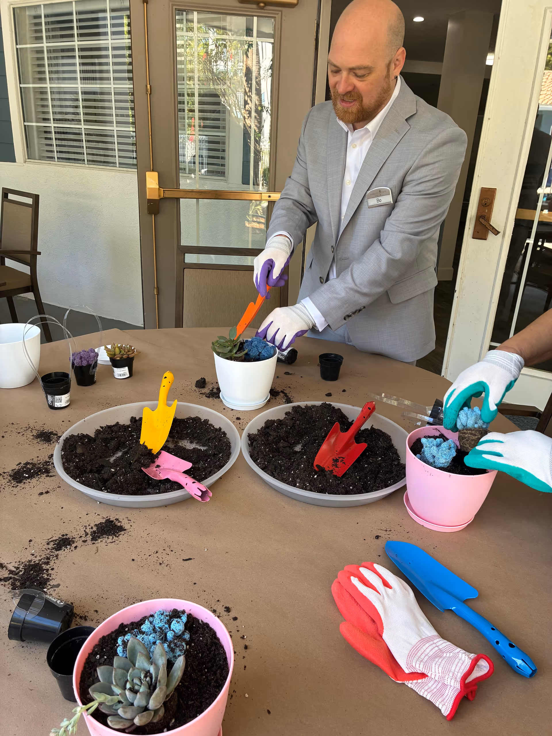 A man in a gray suit pots a plant at an outdoor patio table.