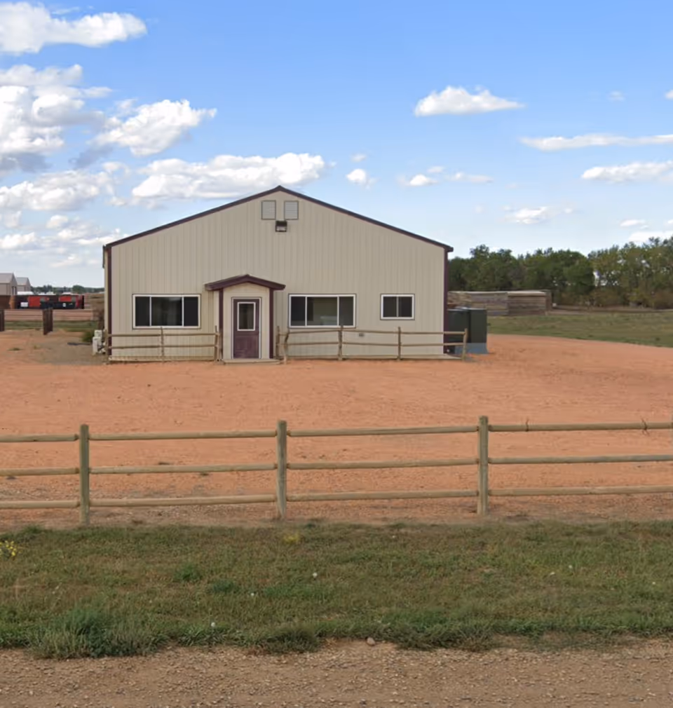 Rural beige metal building with a small porch and wooden fence in a dirt lot.