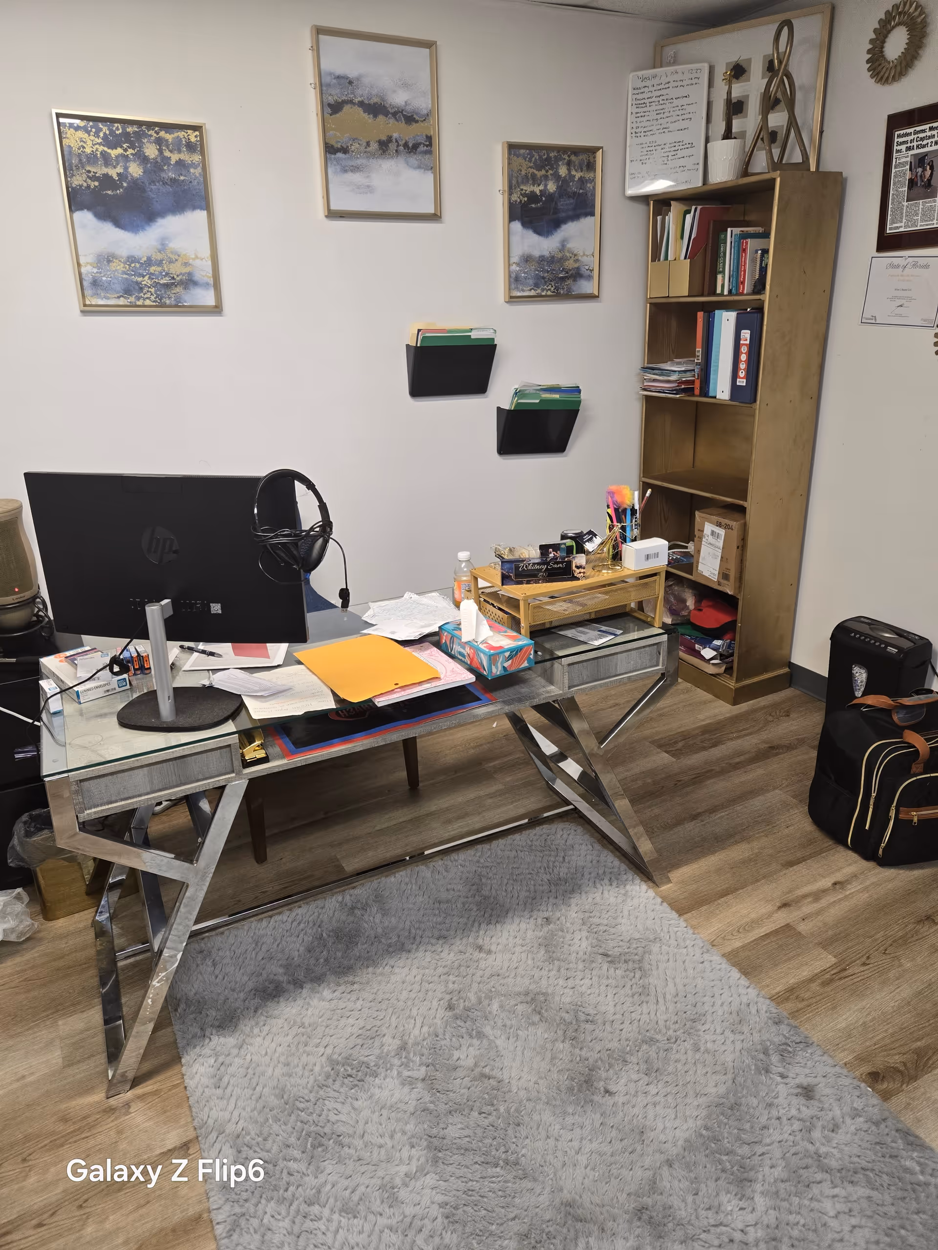 Glass desk with computer, headphones, papers, and a tall bookshelf in an office.