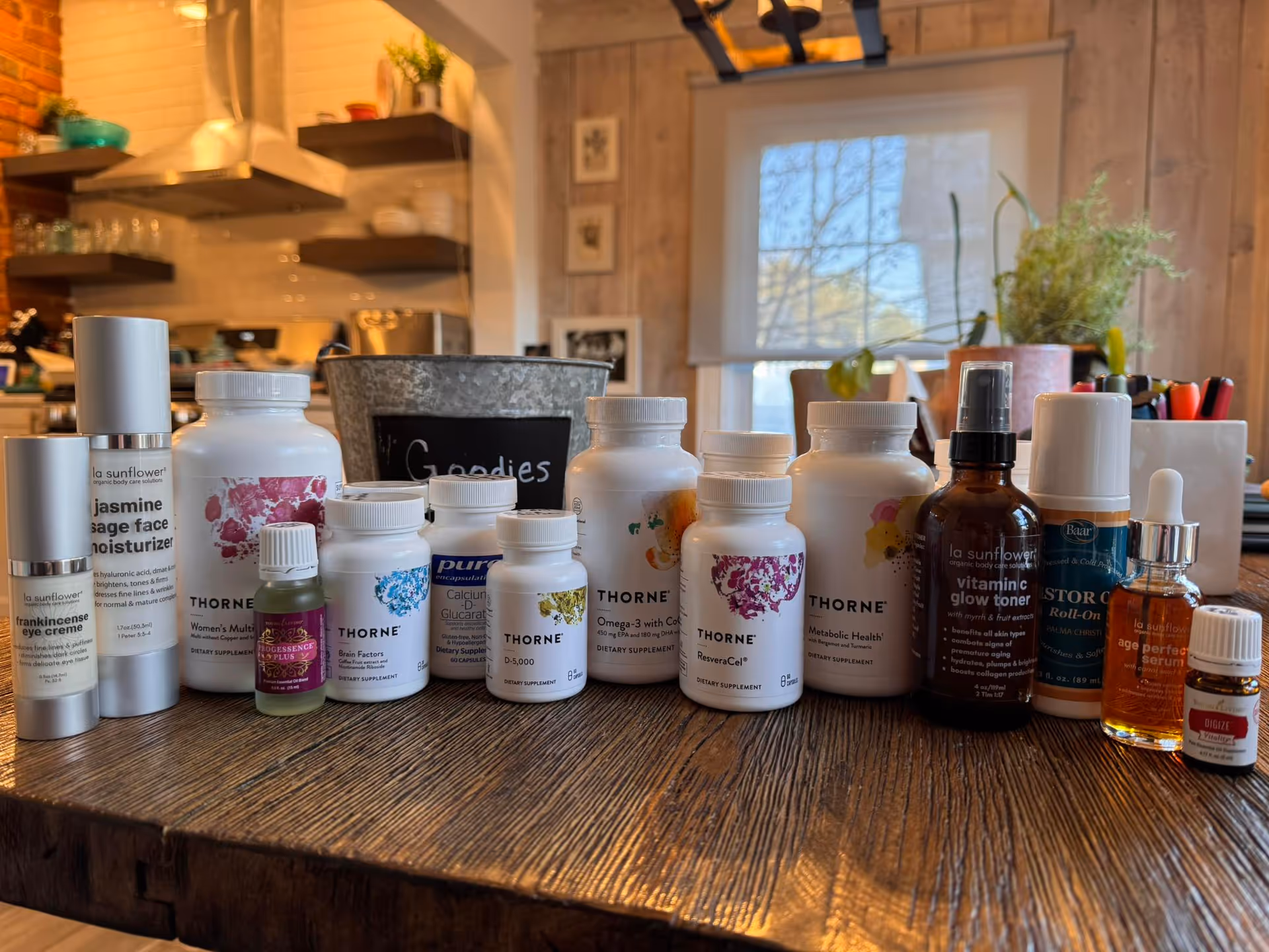 A wooden table is cluttered with vitamin bottles, with a kitchen backdrop and plants.