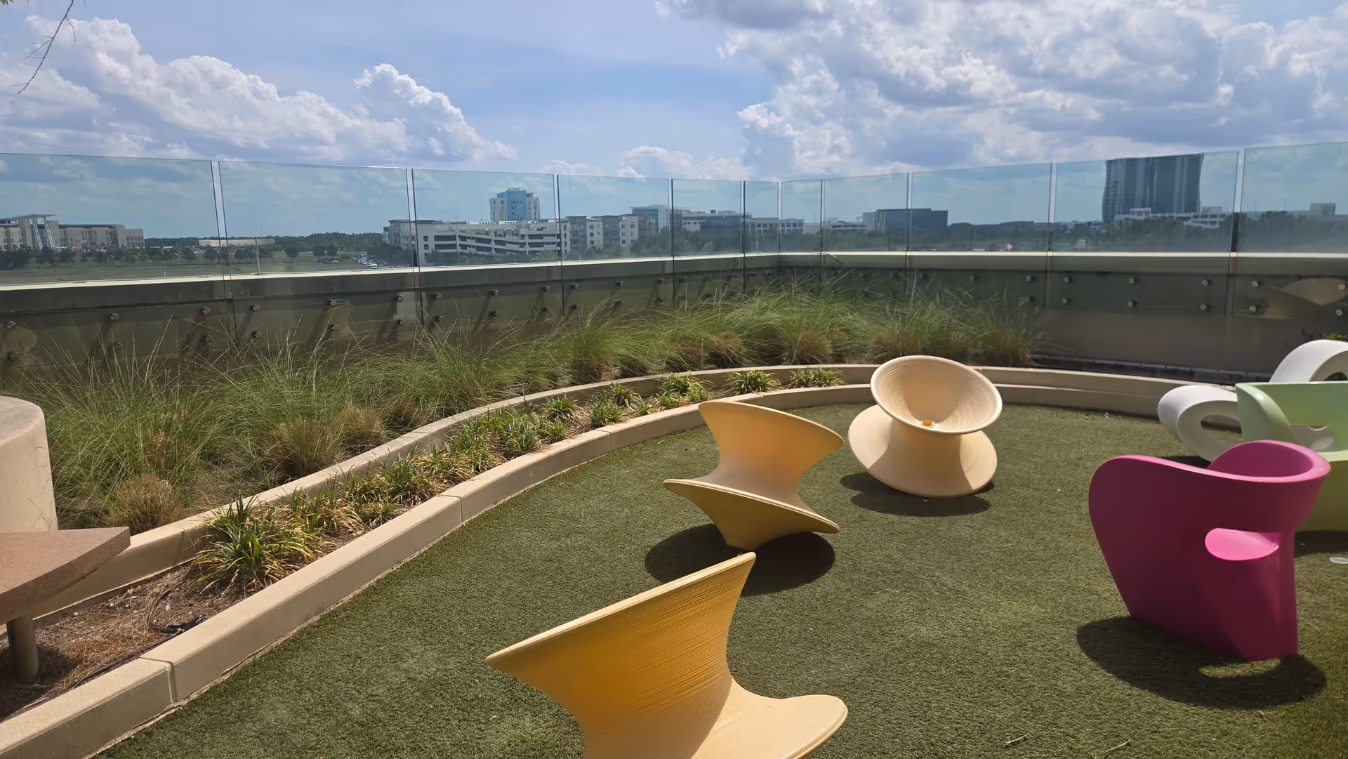 Outdoor rooftop terrace with colorful modern chairs, planters, and glass railing overlooking city skyline.