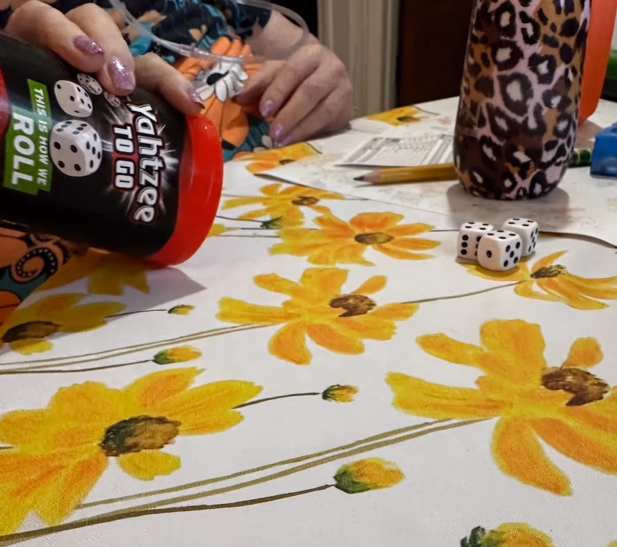Hands and a leopard-pattern cup on a floral tablecloth with dice and pencils.