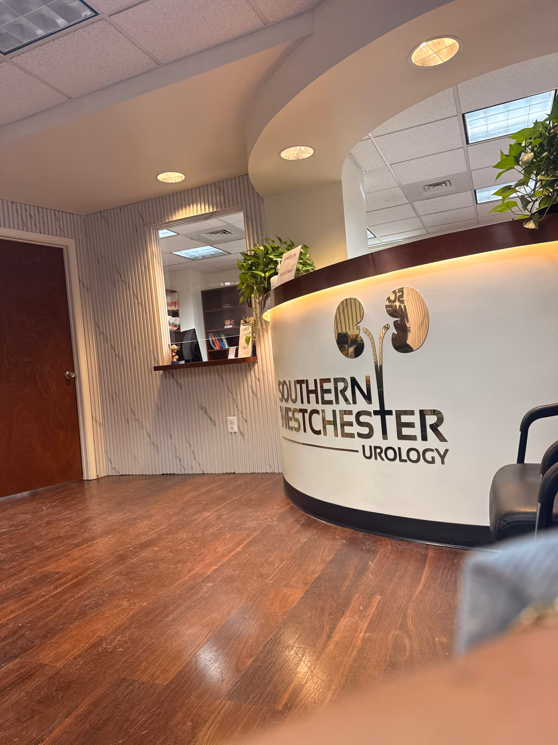 Curved reception desk with Southern Westchester Urology logo, wood floor, plants, and ceiling lights.