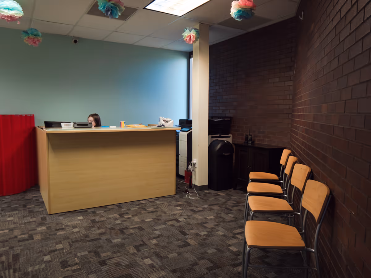 Reception area with a wooden desk, a person, orange chairs, brick wall, hanging pom-poms.