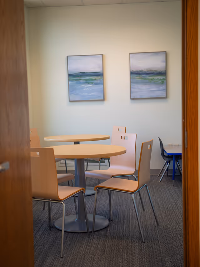 Two round wooden tables with light chairs in a small dining room, wall art above.