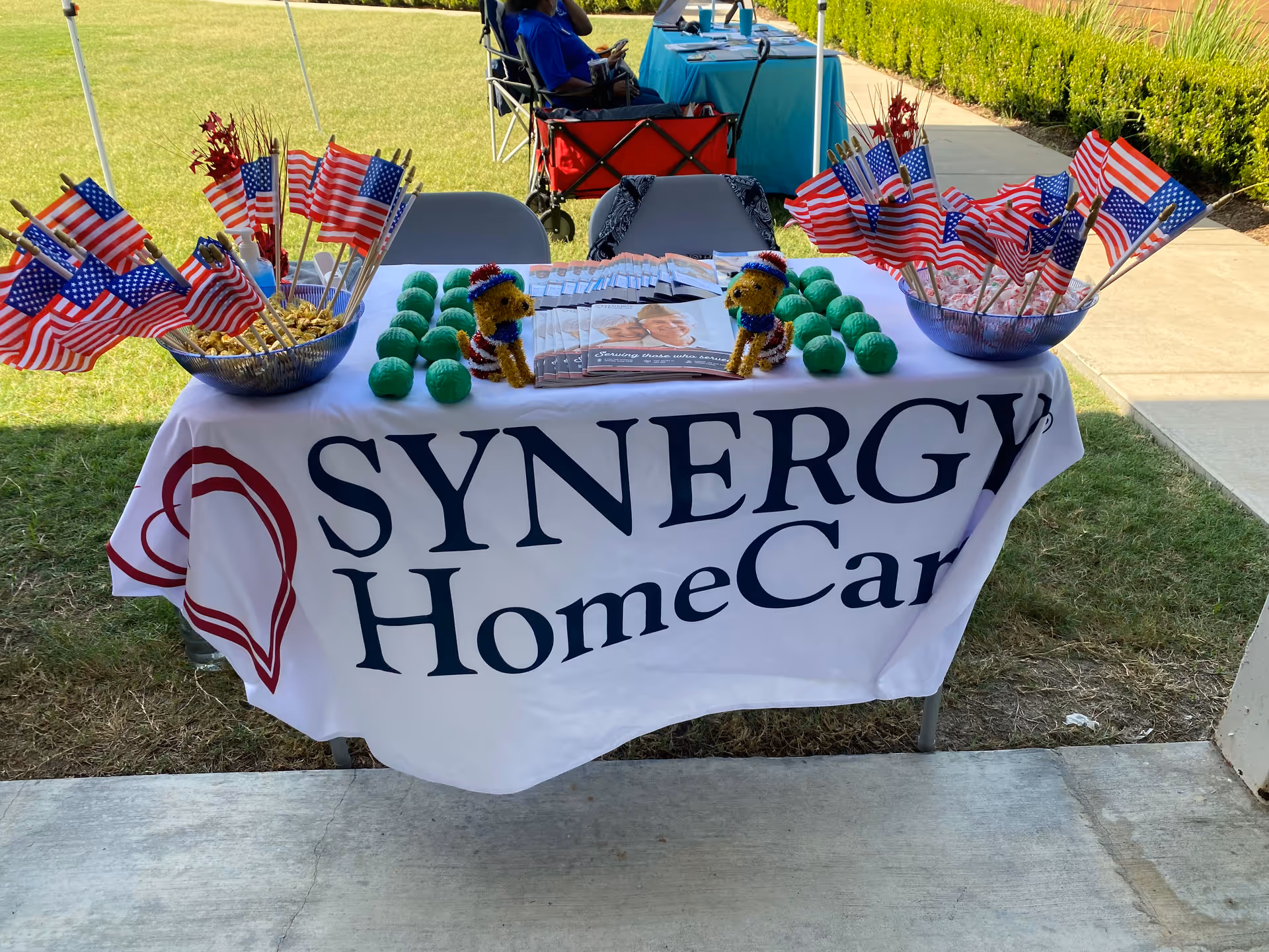 Outdoor event table with SYNERGY HomeCare banner and American flags on lawn.