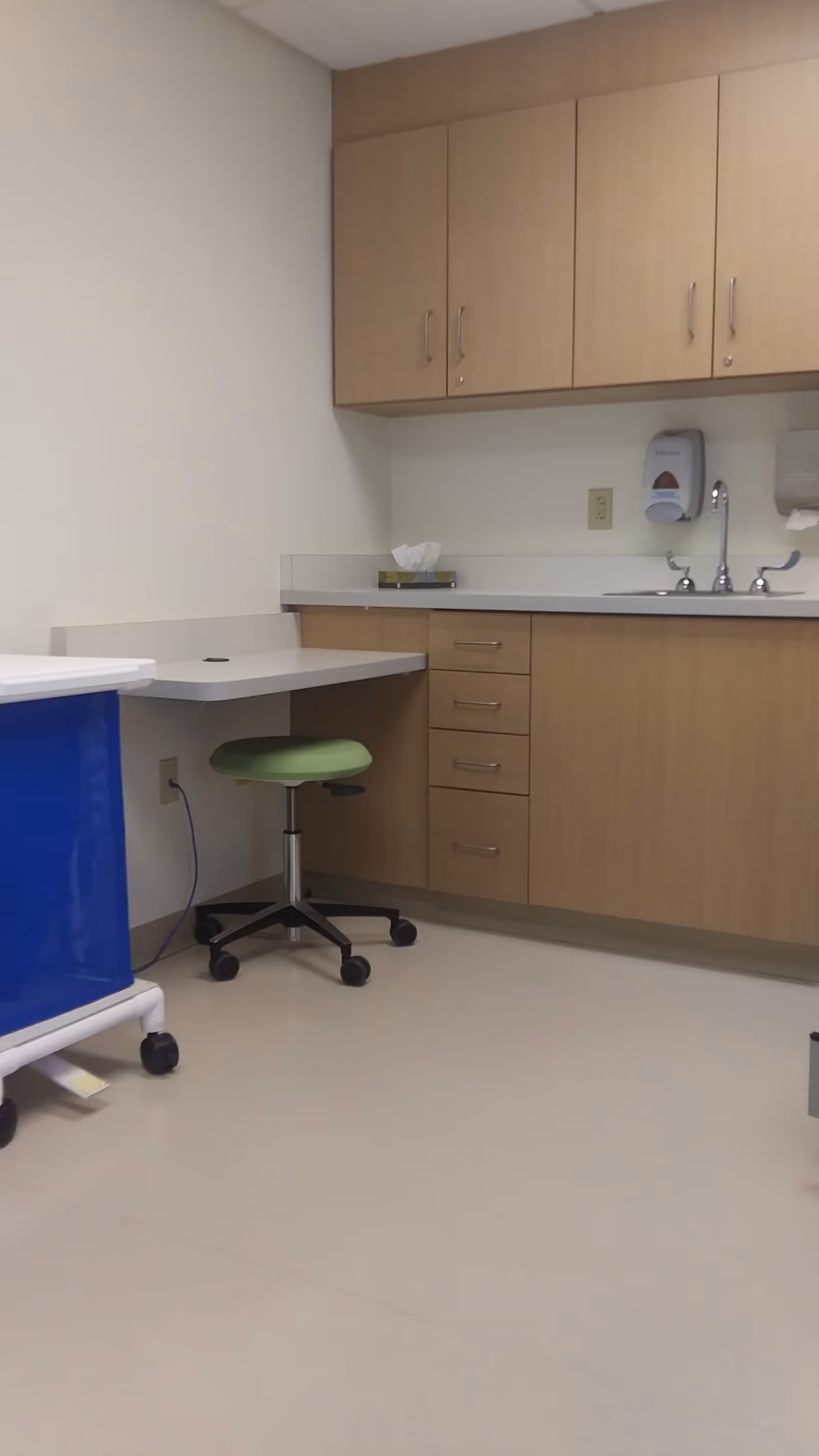 A clean hospital kitchenette with wooden cabinets, a sink, and a green stool.