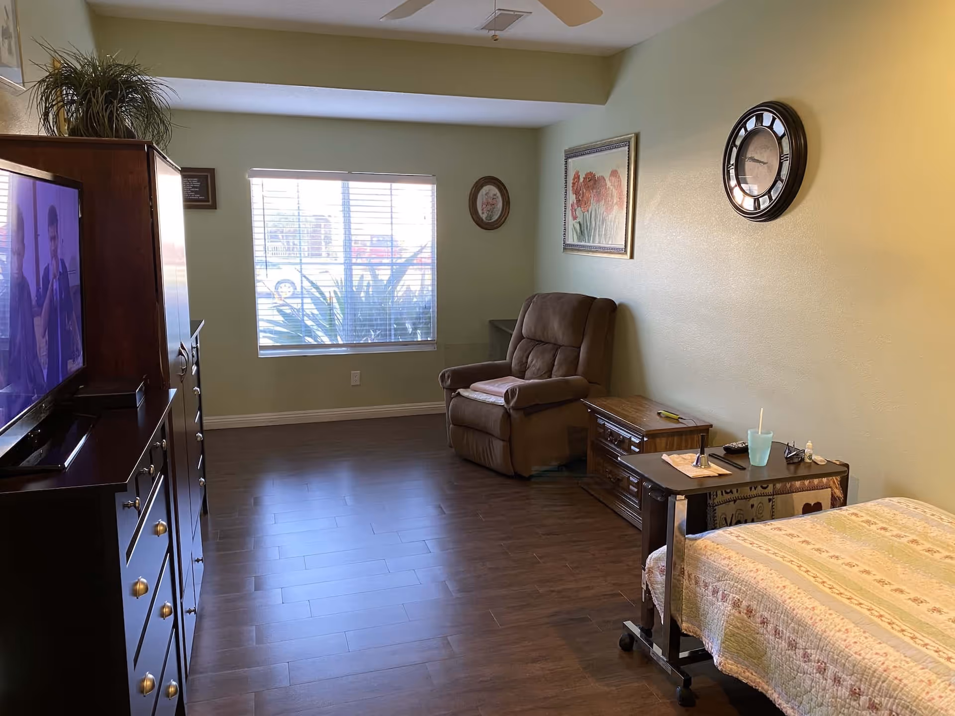 A cozy senior living room with a recliner, TV, dresser, and bed alongside a window.