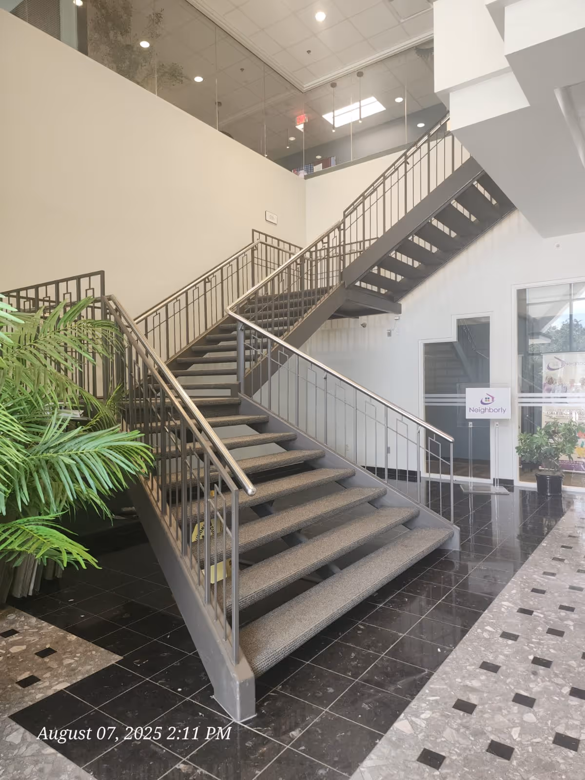 Indoor stairwell with metal railings, gray carpeted steps, white walls, and potted plants.