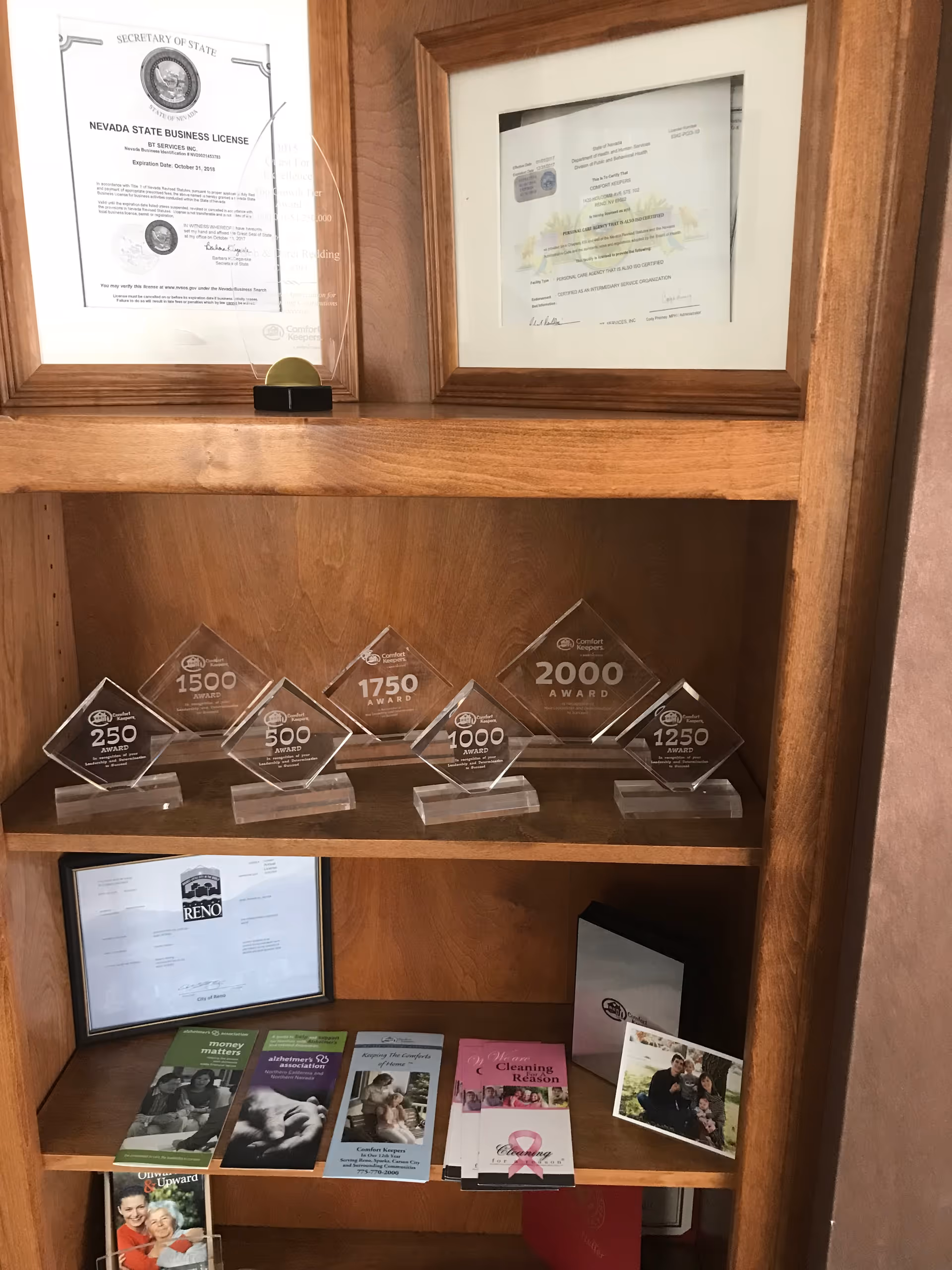 Wooden display shelf with framed licenses, glass awards, and brochures inside a senior care facility.