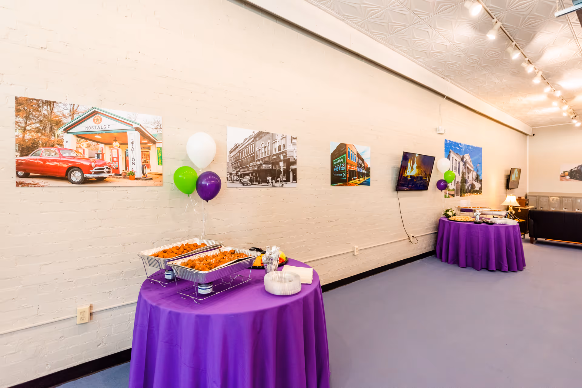 Indoor event space with purple tablecloths, food trays, balloons, and wall art.