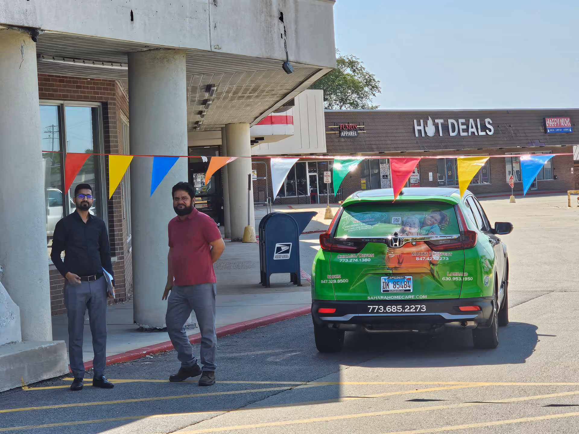 Two men stand outside Sahara Home Care entrance under colorful pennant flags