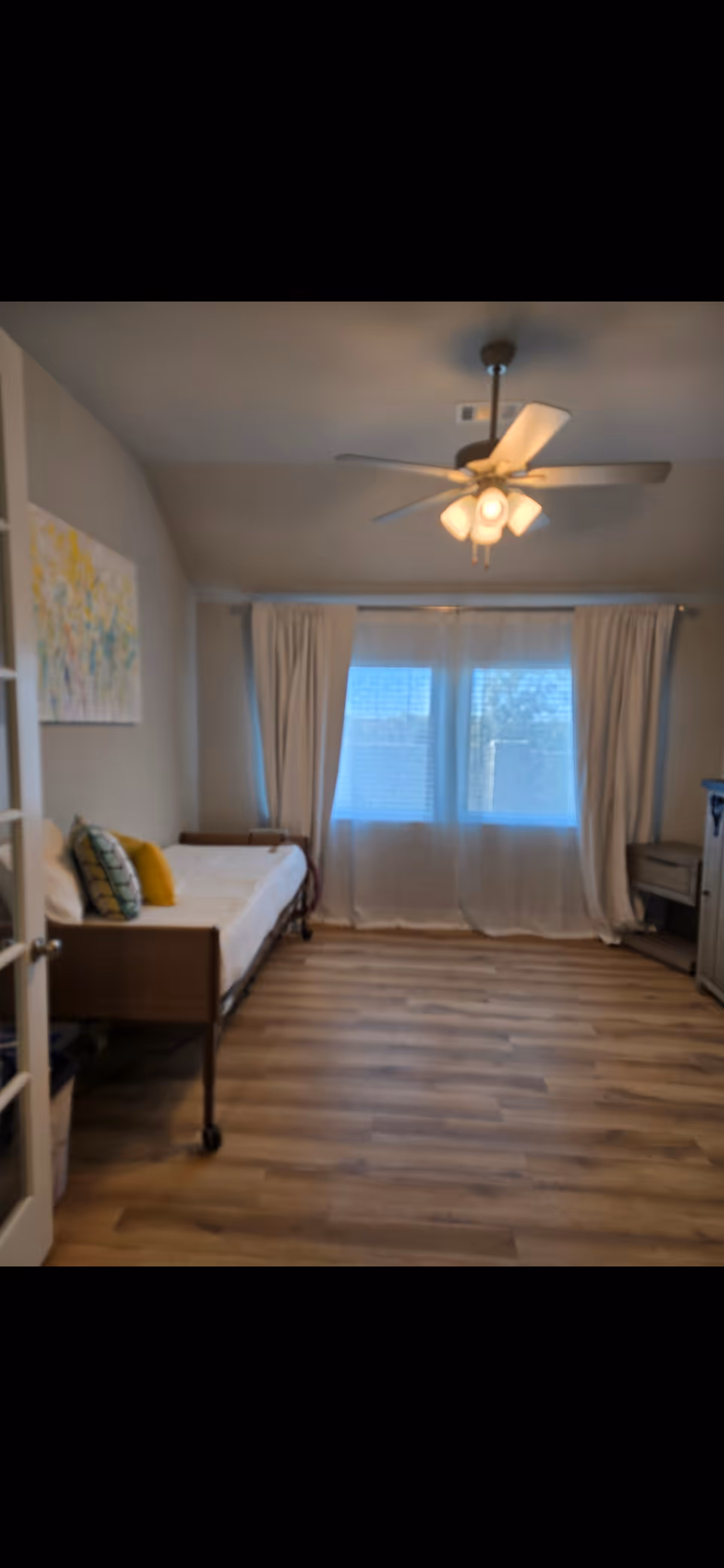 A sparsely furnished senior living bedroom featuring a bed, windows with curtains, and a ceiling fan.