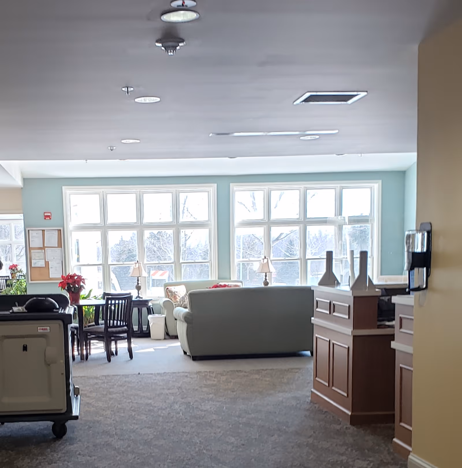 A bright hospice lobby with large windows, teal walls, and sofas, chairs, and a reception desk.