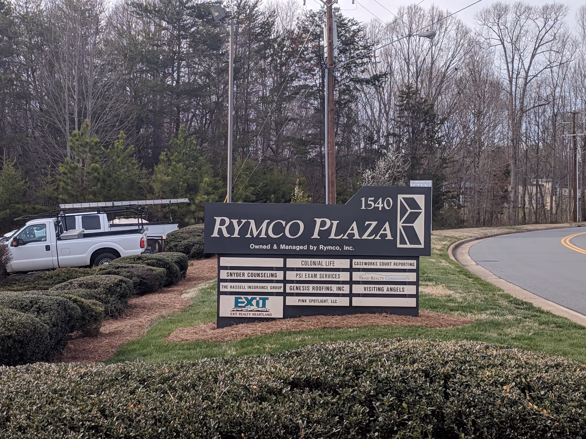 Exterior view of a plaza sign reading RYMCO PLAZA, with shrubs and a white pickup truck.