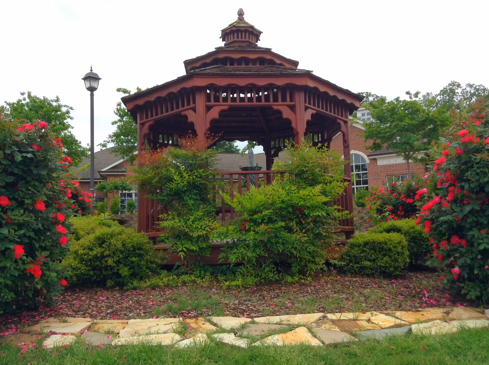 Wooden gazebo in a garden with red flowers and a brick building in background.