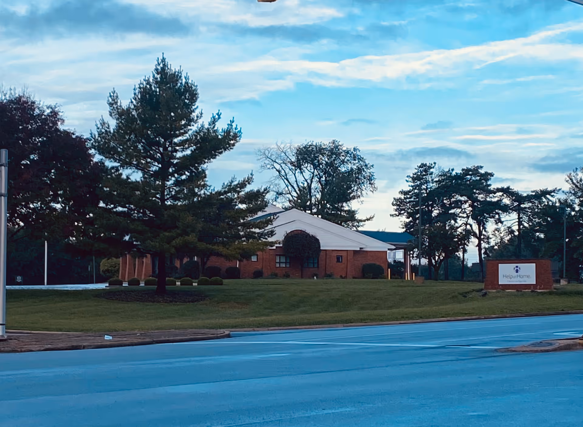 Brick senior living facility with white roof, trees, and a Help at Home sign.
