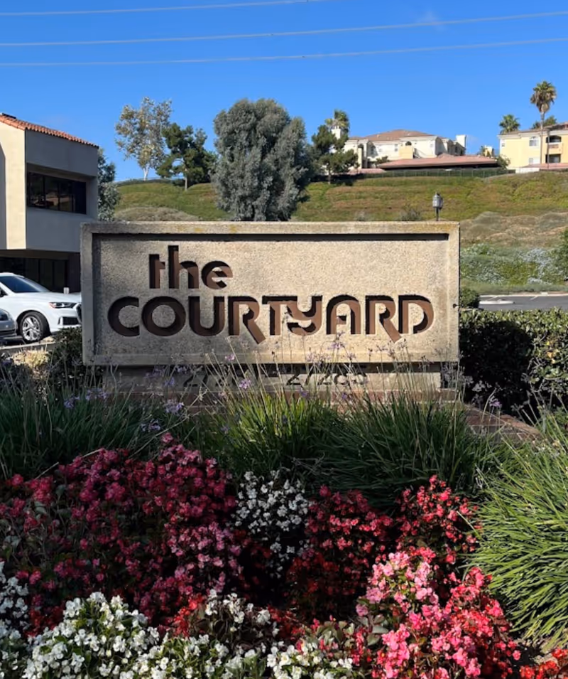 Outdoor exterior scene featuring a stone sign reading 'the courtyard' with colorful flowers in front.