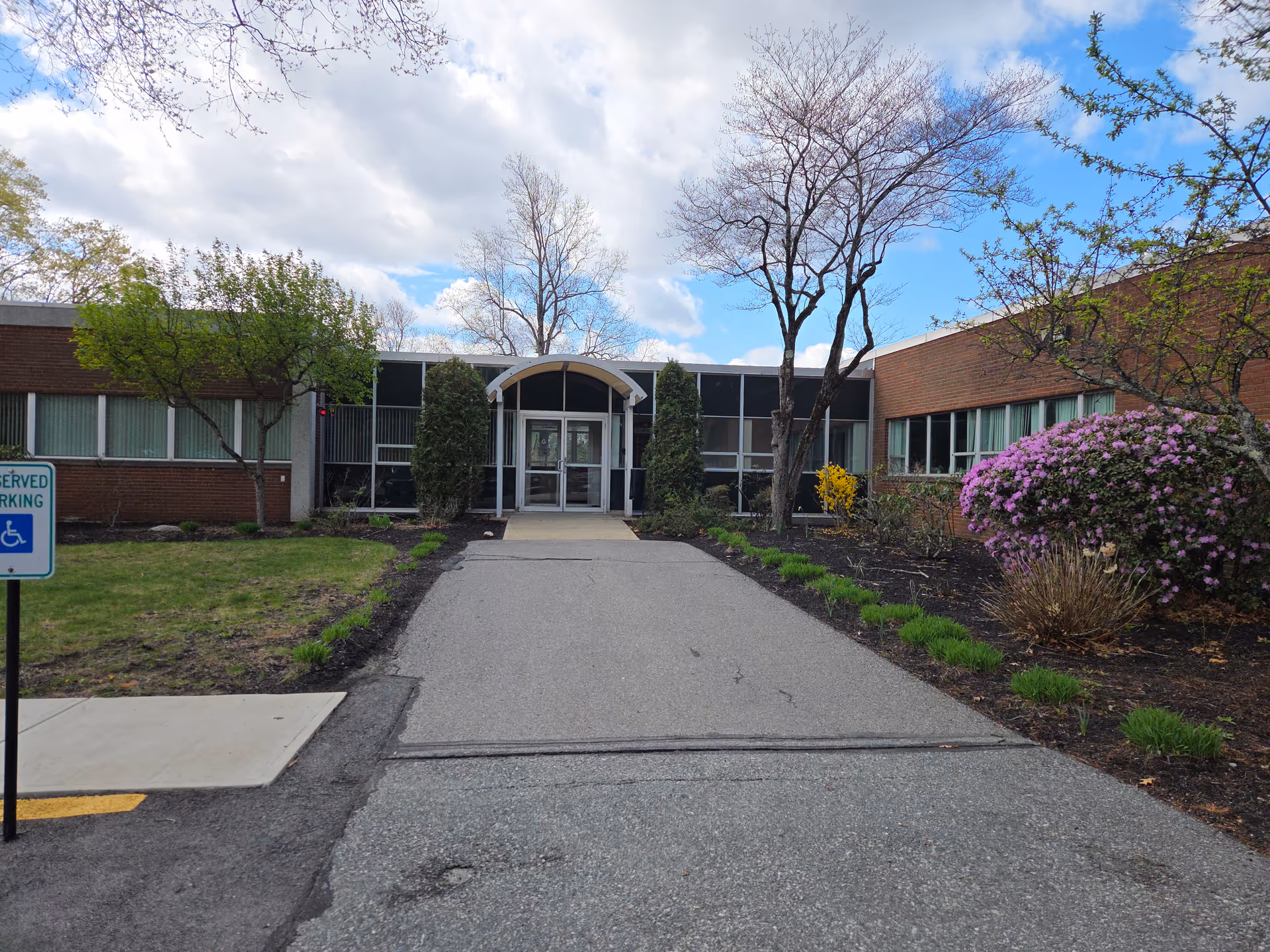 Paved walkway leads to glass-front entrance of a low-rise building with greenery.