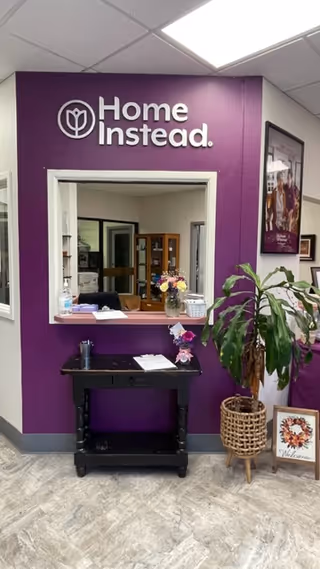 Reception area with purple Home Instead wall, pass-through window, plant, and framed art.