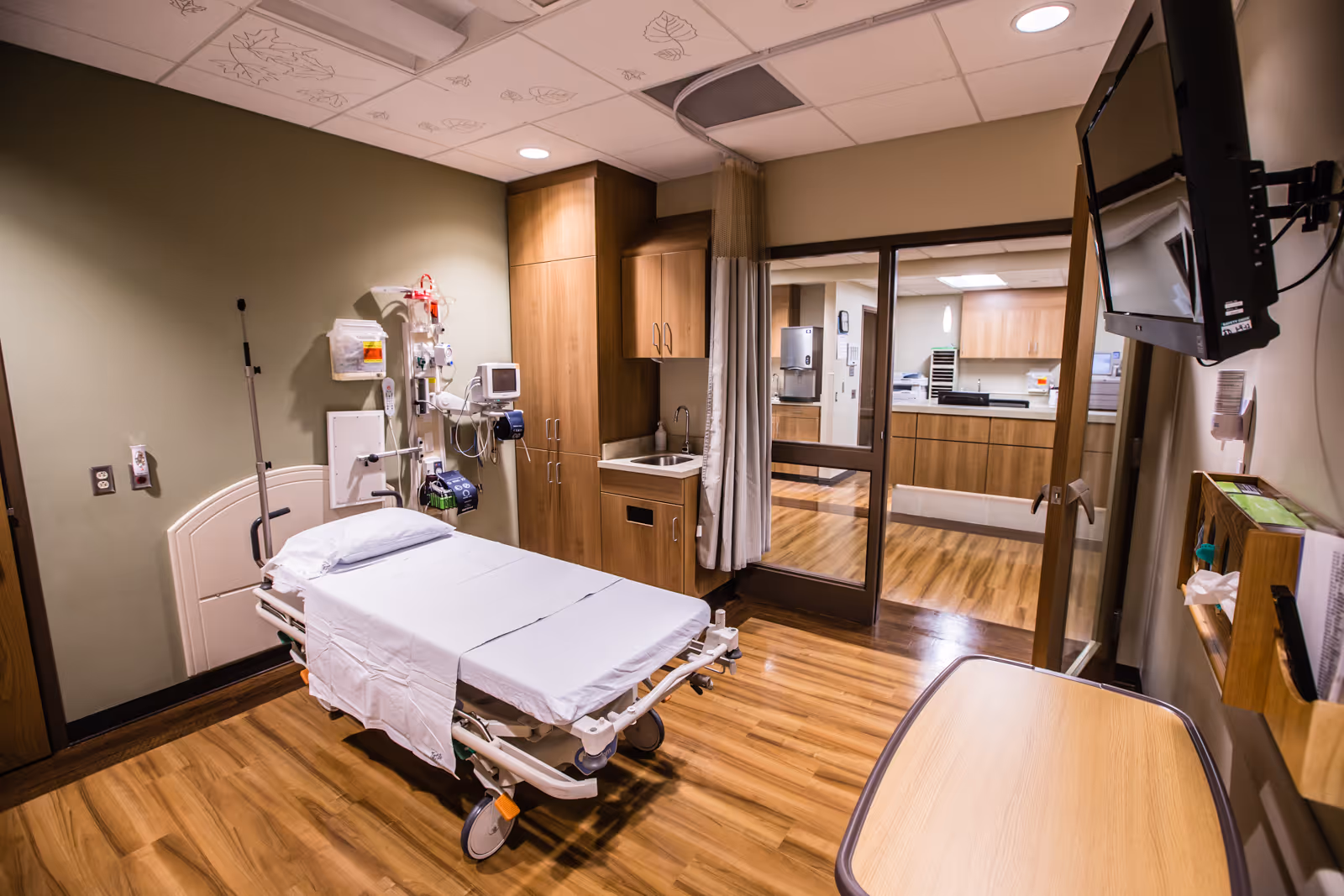 Hospital-style patient room with adjustable bed, medical equipment, cabinets, and doorway to hallway.